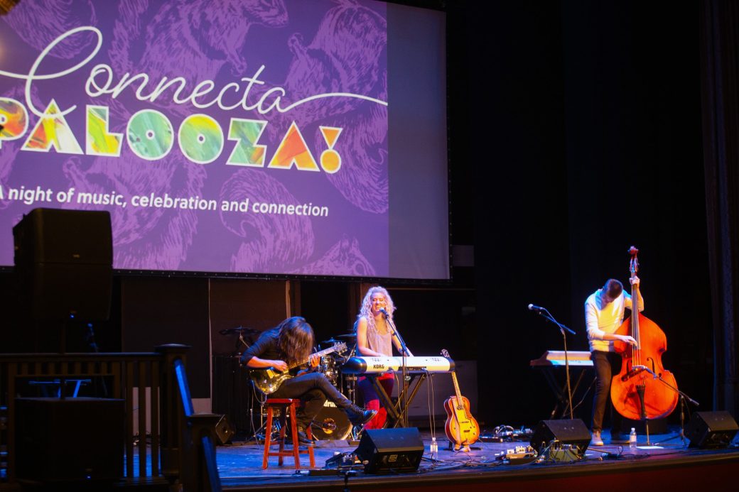 Woman singing at Conctpalooza and playing acoustic guitar and two men playing bass