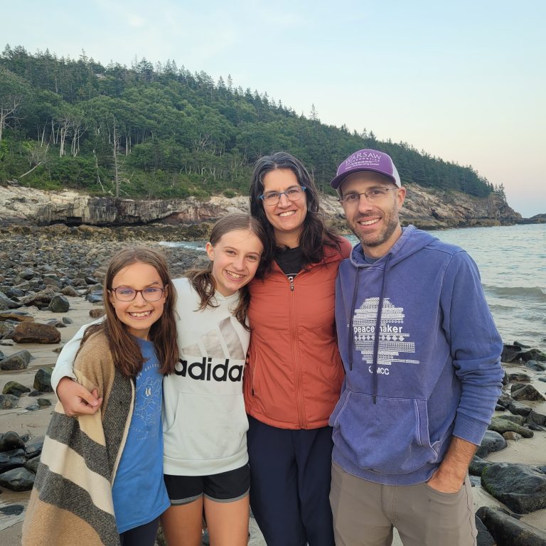 A family of two adults and two children stands together on a rocky beach.