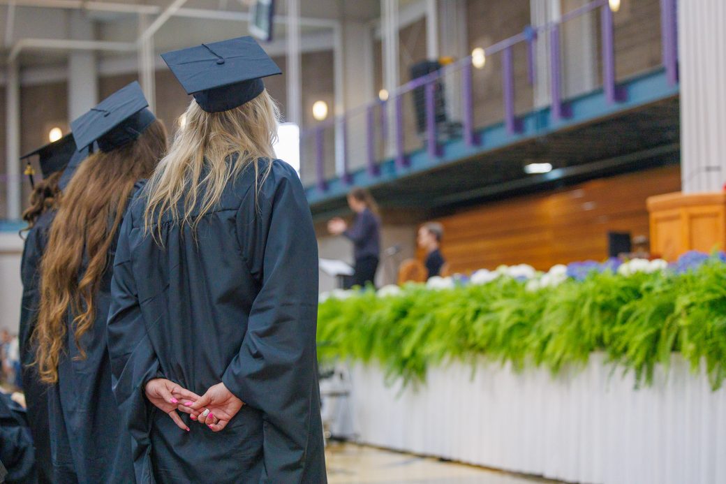 Graduate with hands clasped behind back looking toward stage