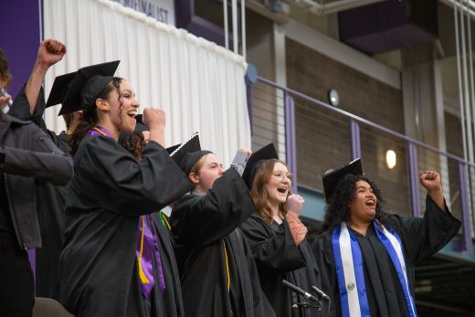 students stand on stage in graduation caps and gowns celebrating