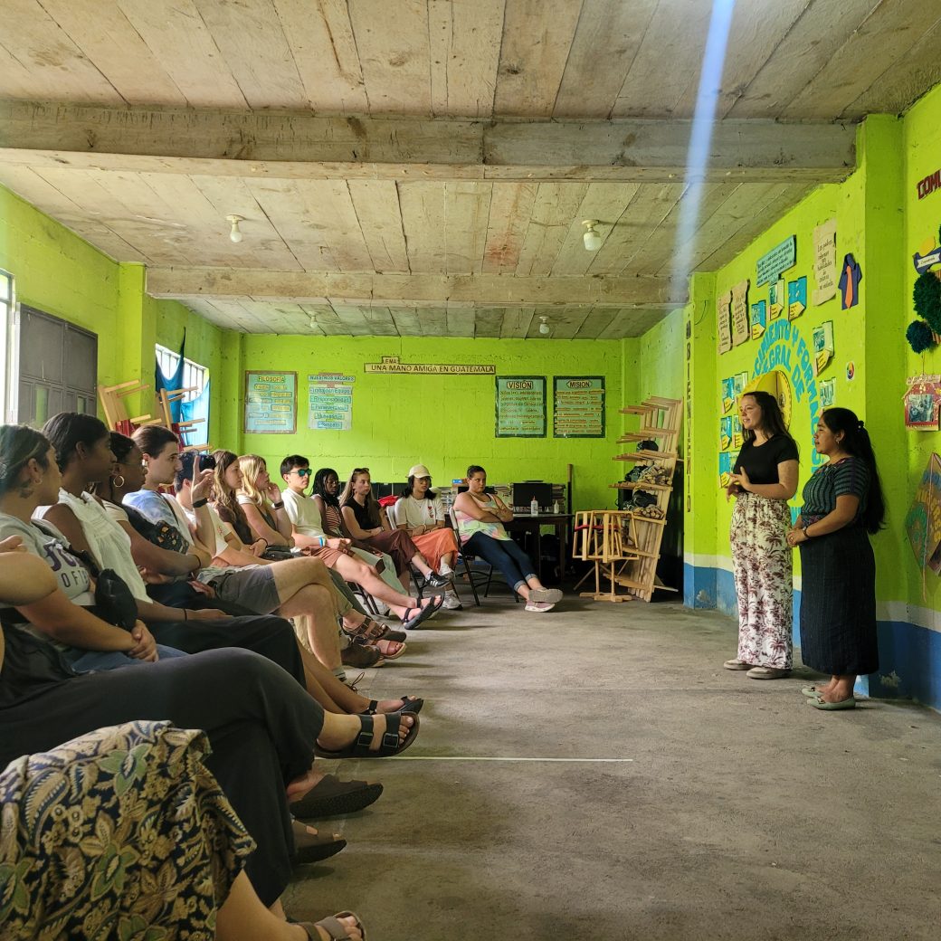 A large group of people seated in a lime green room, listening to two speakers.