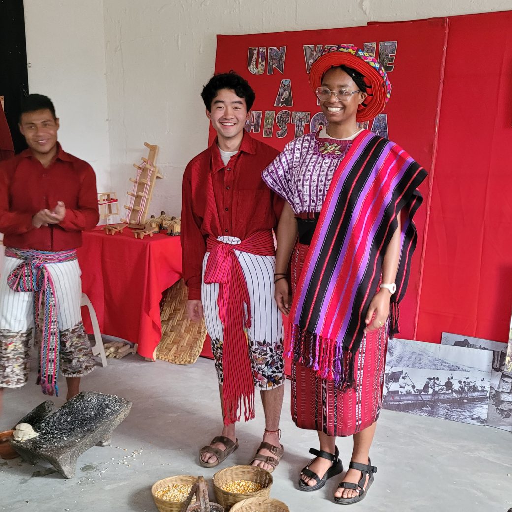 Two people stand smiling, while wearing colorful red, white and purple clothing.