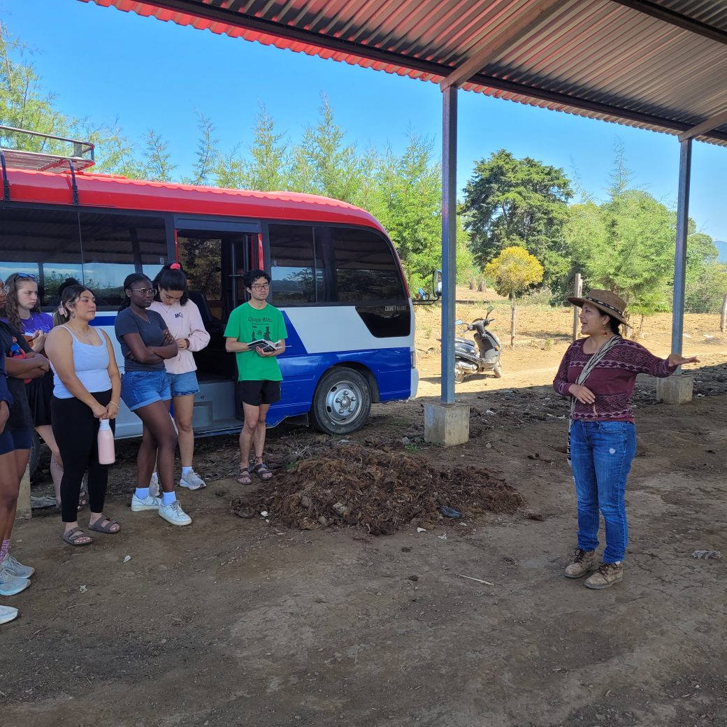 A group of people in front of a bus, listening to a speaker.