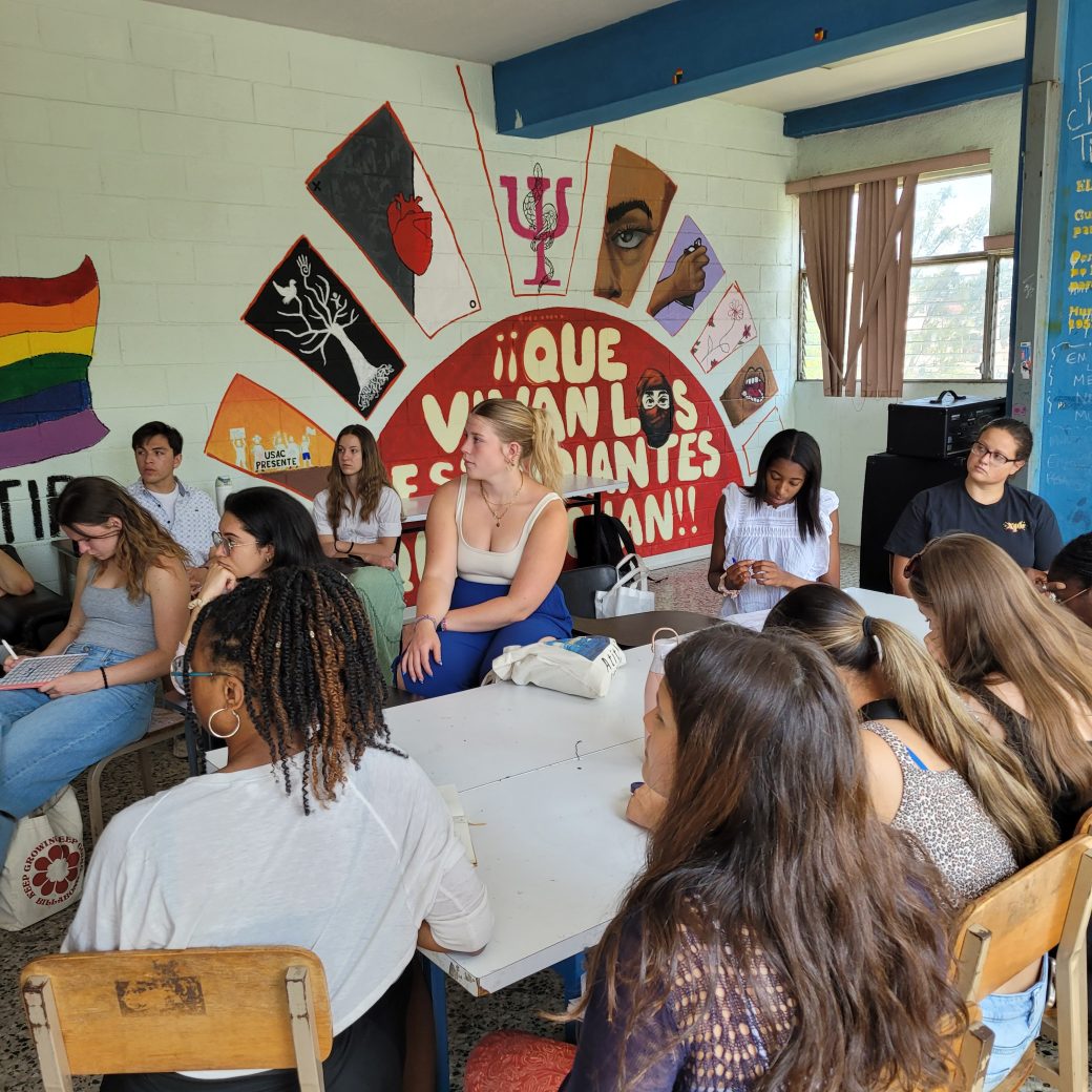 A group of people seated around a table, listening to a lecture.