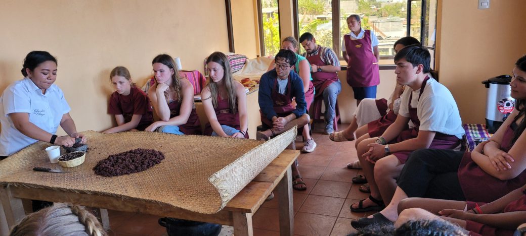 A large group of people in a small room, gathered around a table with a mound of ground chocolate in the center.