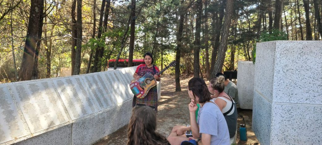 A woman with a guitar standing next to a group of people and a white stone wall.
