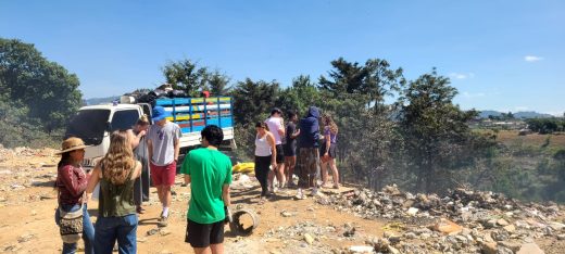 A group of people in a dusty landscape, standing in front of a truck filled with trash bags.