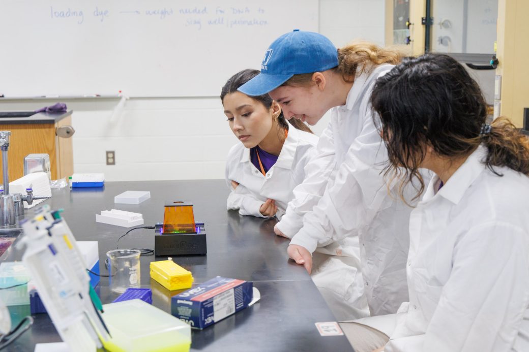 Three high school students look at a lab test in white coats for summer camp
