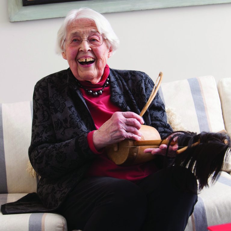Mayer Oyer in her living room playing a traditional African instrument and laughing at someone behind the camera