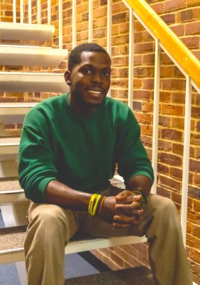 A young man in a green sweater sits on a brick staircase, smiling slightly with his hands clasped.