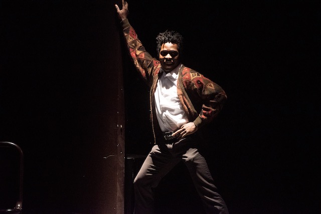 A student performer stands iin a disco pose on stage wearing a brown-red cardigan.