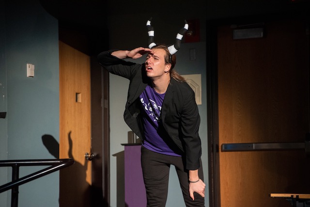 A student performer representing Evil wearing a purple GC shirt ,a black sport coat and with striped horns, peers into the crowd while shading his eyes from the stage