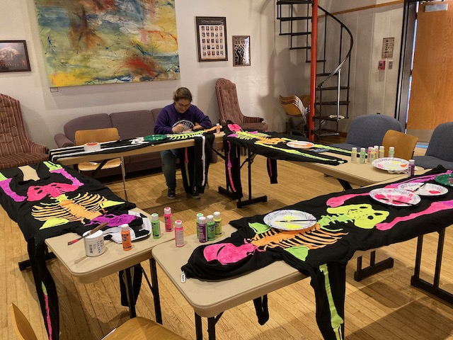 A single person sits at a large square of long tables covered in the painted and drying skeleton costumes.