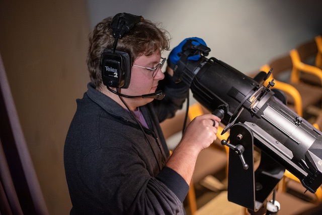 A studnet wearing dark clothing wearing headphones stands behind a spot light looking down toward the stage