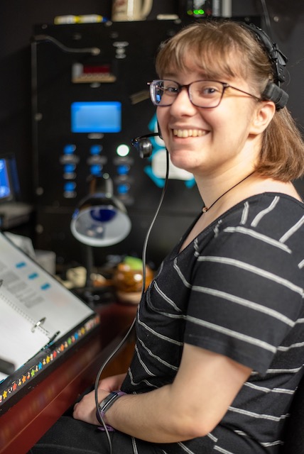 A student wearing a black shirt with this white stripes, glasses and a headset smiles at the camera