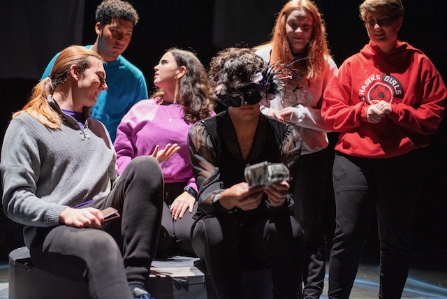 A group of student performers in colorful tops and dark pants is gathered on stage facing the aduience but interacting with each other.