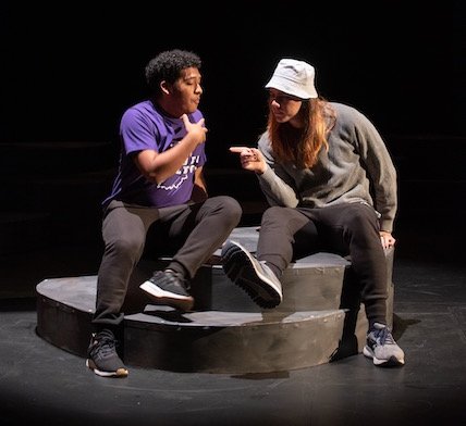Two student performers onstage sitting on a prop in conversation.
