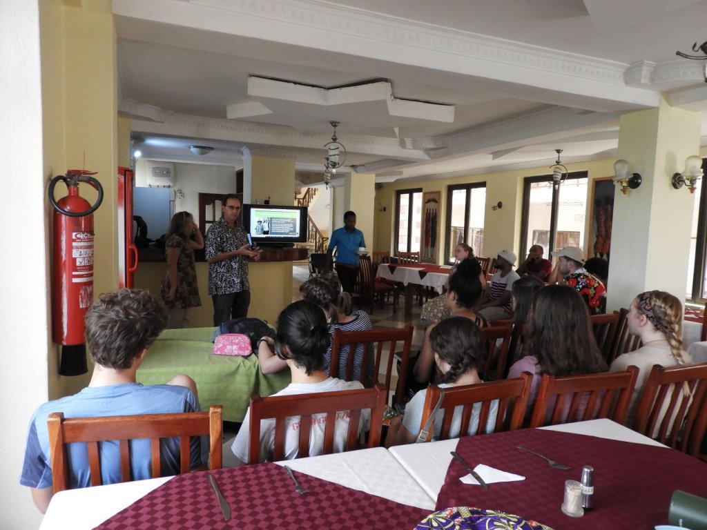 People seated at tables watching a projected slideshow presentation in a room.