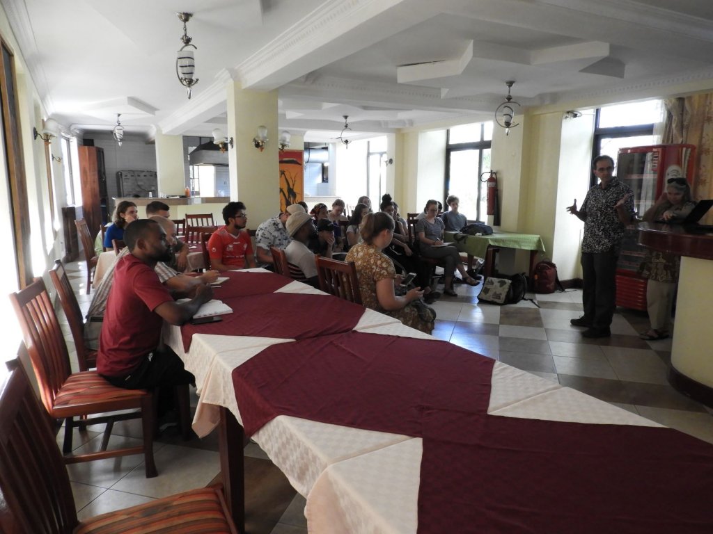 People seated at long tables listening to a speaker standing at the front of a room.