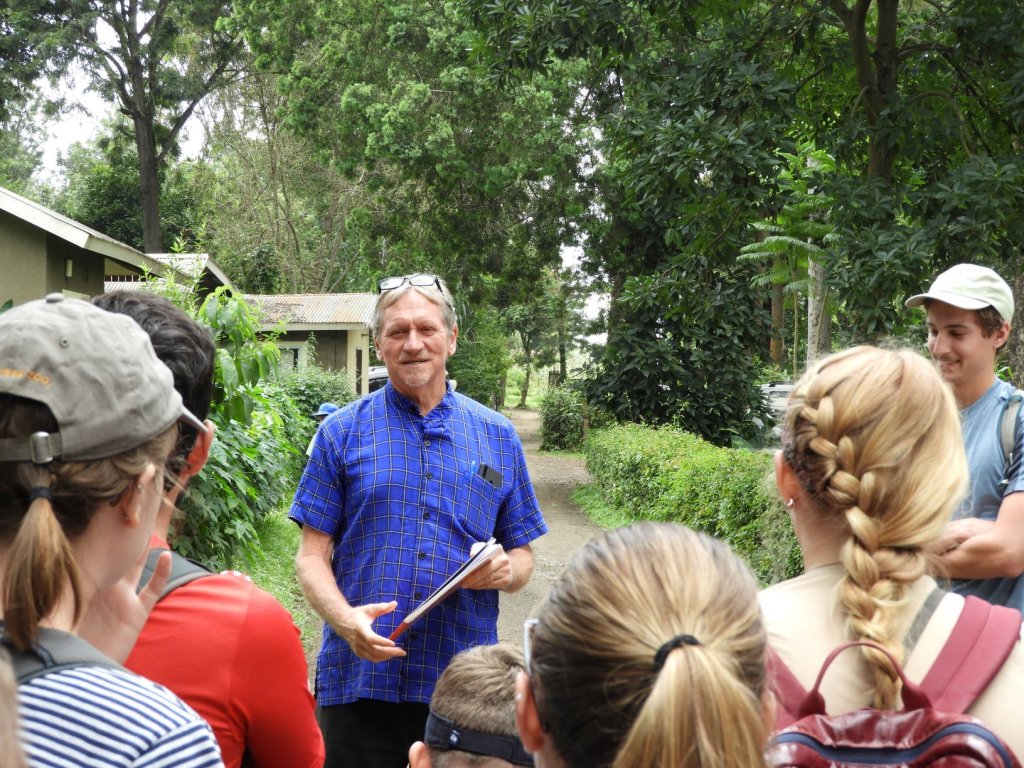 A man in a blue shirt holding a clipboard speaks to a group of students gathered around him outdoors.