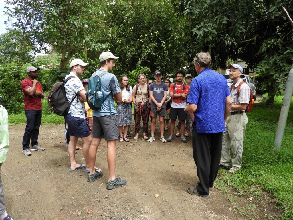 A group of students gathered on a dirt path outdoors, listening to a guide during a tour.