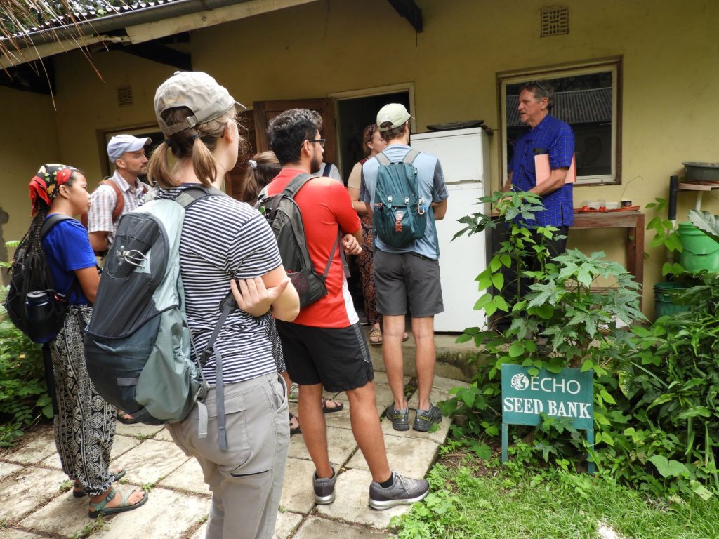 A group of students standing beside an ECHO Seed Bank sign, listening to a presenter outside a building.