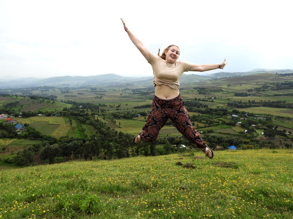 A woman jumps with her arms outstretched on a grassy hilltop overlooking a scenic green valley.