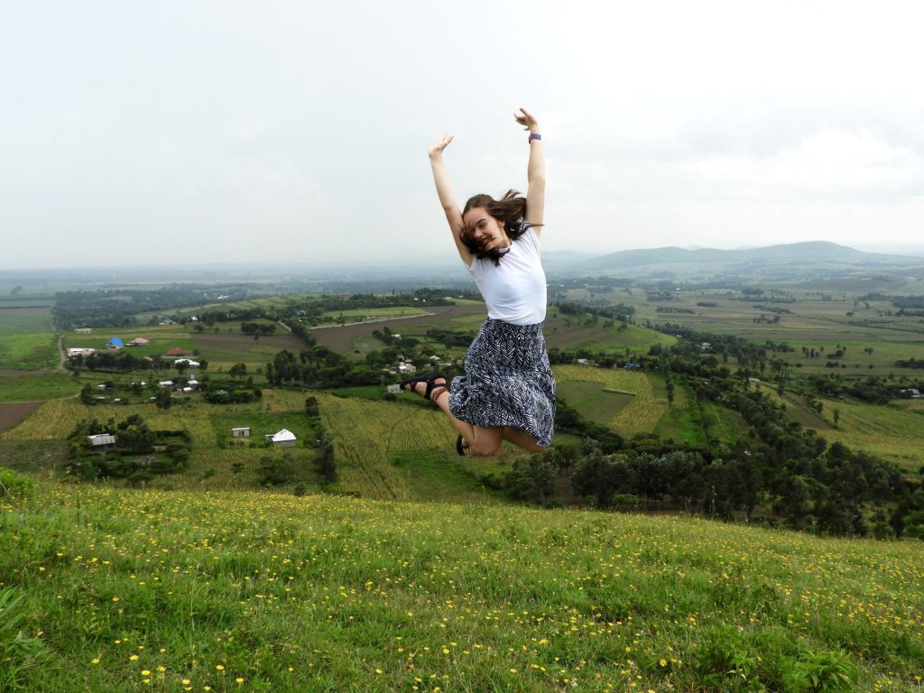 A woman leaps into the air with her arms raised on a grassy hill overlooking a lush, rolling green landscape.