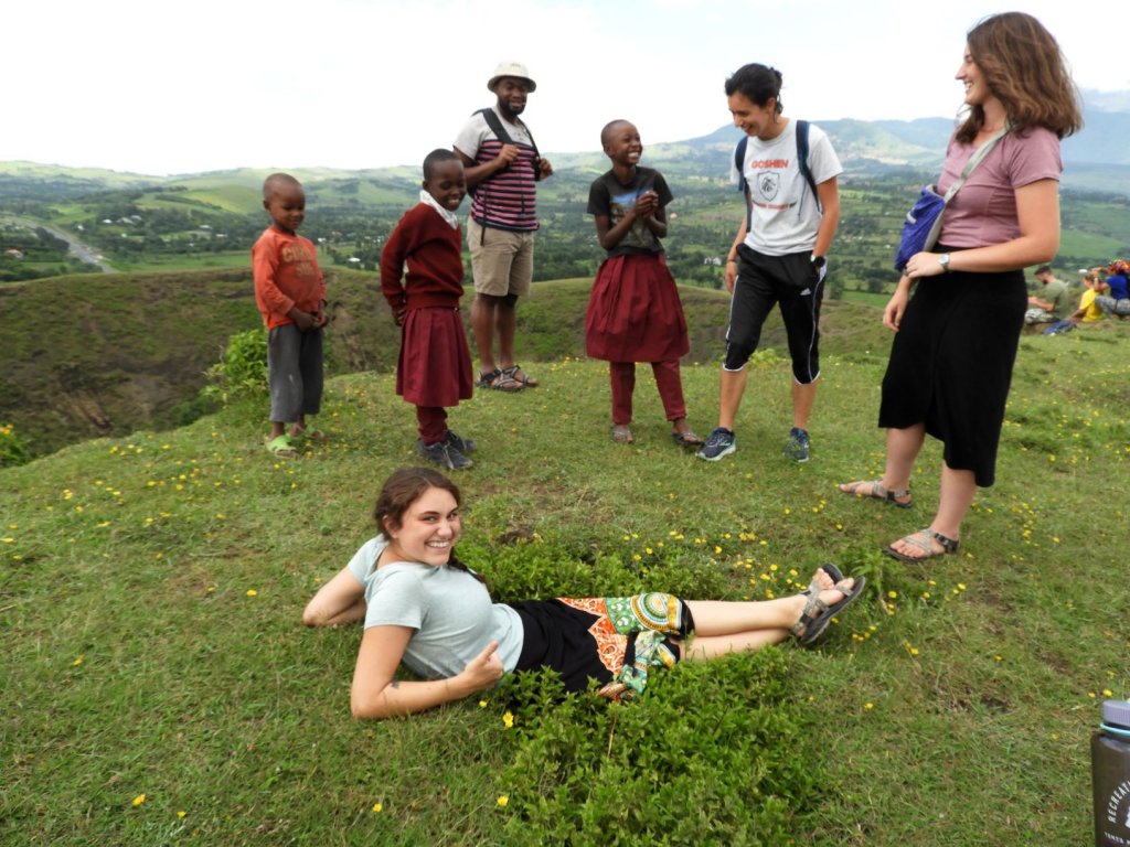 A group of students and local children stand together on a grassy hillside in the Serengeti while a young woman relaxes on the grass in the foreground.