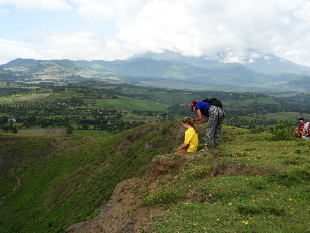 Two people stand on a grassy ridge overlooking a vast green valley of farmland with mountains rising in the distance.