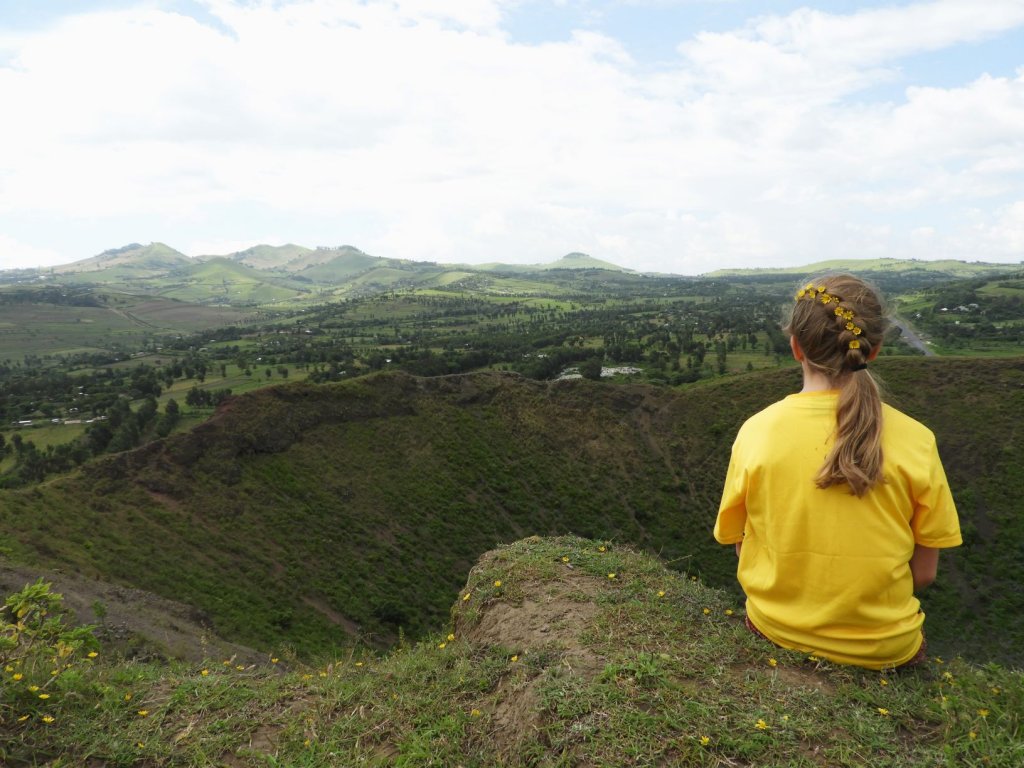 A person in a yellow shirt sits on a hilltop seen from behind, gazing out over a green valley and rolling hills under a cloudy sky.