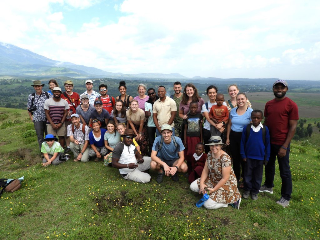 A large group of students and local community members, including several children, pose together on a grassy hilltop with mountains and a valley in the background.