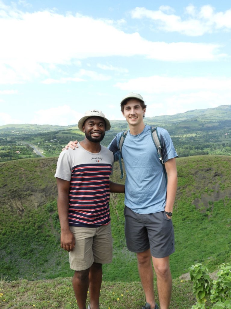 Two smiling young men stand side by side with arms around each other's shoulders on a green hillside overlooking a valley.