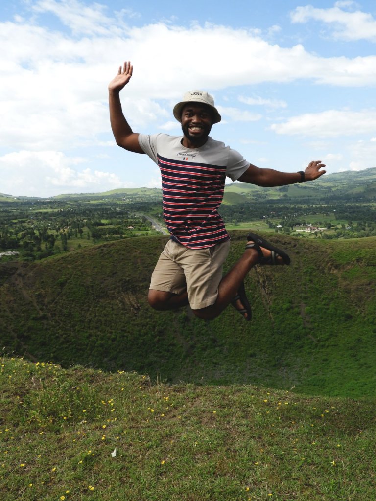 A man in a striped shirt and hat jumps joyfully into the air on a grassy hilltop overlooking the Serengeti landscape.