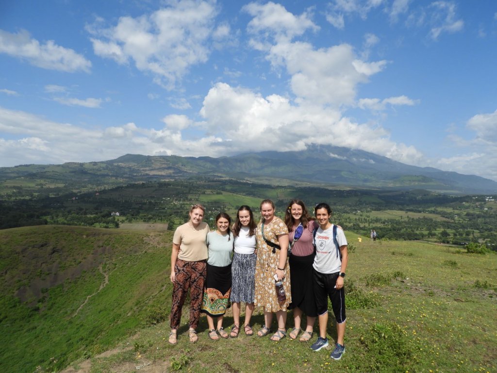 A group of students posing together on a green hillside with mountains and clouds in the background.