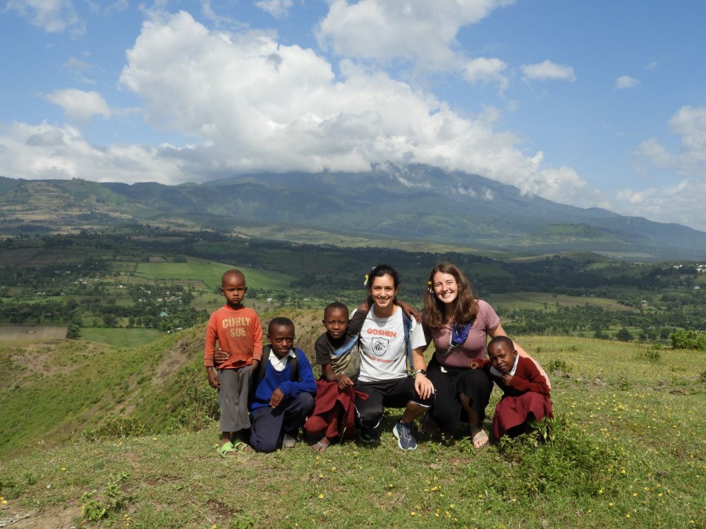 A group of students posing with several local children on a grassy hillside with mountains in the background.