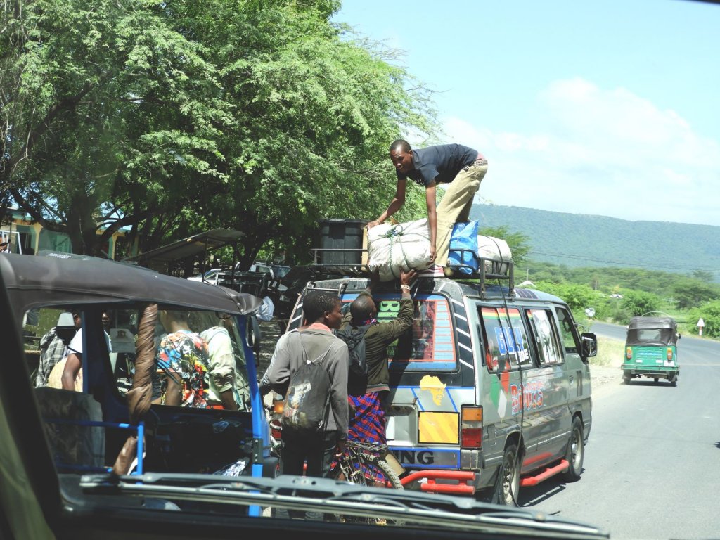 People load luggage and bags onto the roof rack of a minibus parked along a roadside as another vehicle passes by.