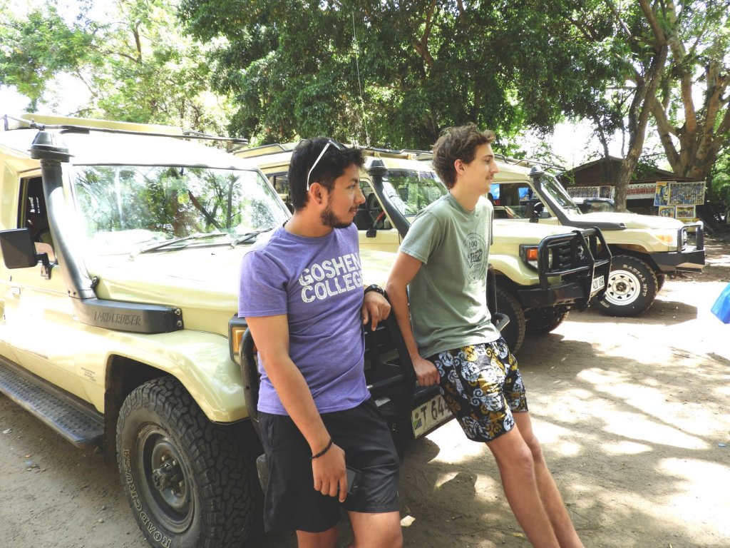 Two young men, one in a purple Goshen College t-shirt, stand beside yellow safari Land Cruisers.