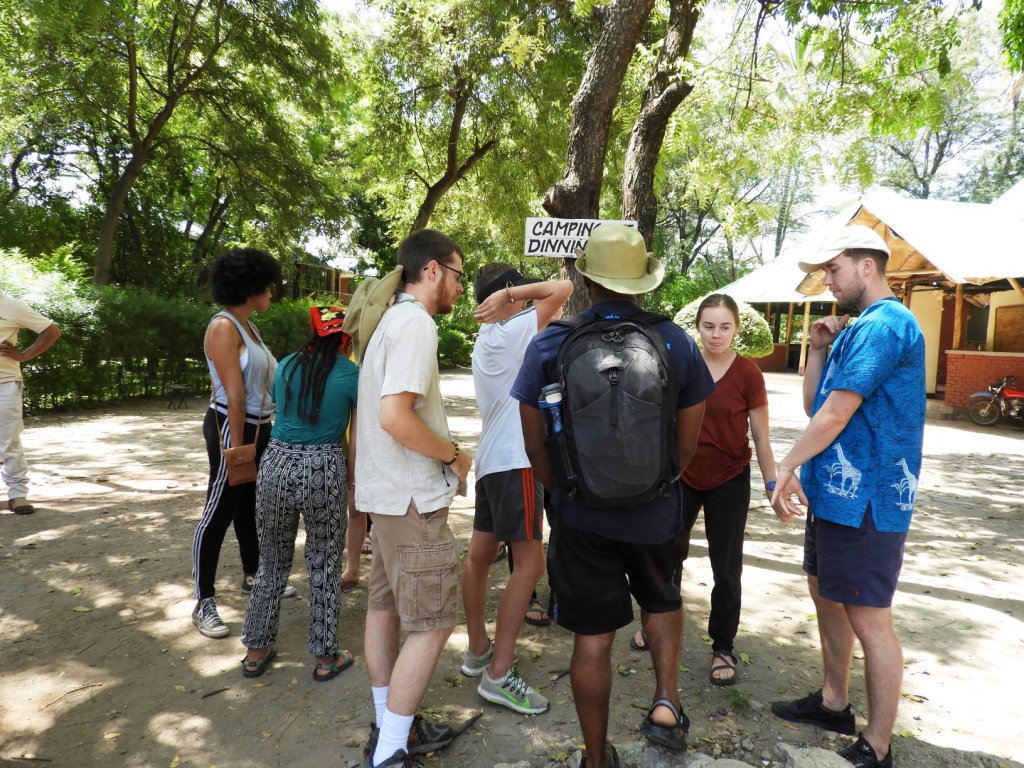 A group of students gathers in a circle at a shaded campsite near a dining tent.