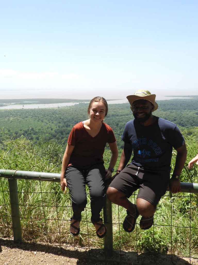 A woman and a man sit on a wooden railing at a scenic overlook with a river and green landscape behind them.