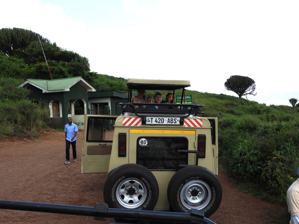A yellow safari 4x4 vehicle parked on a dirt track with a person standing beside it and a green-roofed building nearby.