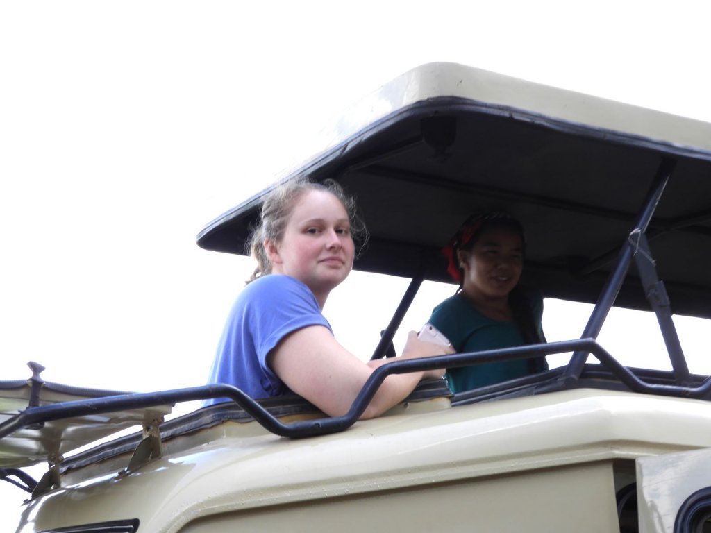 A young woman in a blue shirt sits inside an open-topped safari vehicle with another passenger beside her.