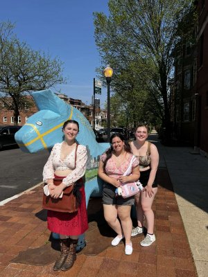 Three young women pose on a sunny sidewalk next to a large blue Swedish Dala horse, one wearing a traditional folk costume with a red skirt and apron.