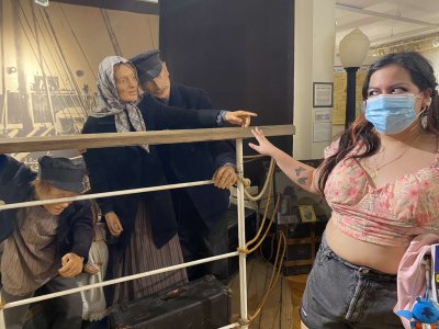 A young woman in a face mask and pink top points toward museum figures dressed in traditional old-fashioned clothing at a heritage exhibit.