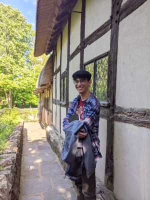 Young man leaning against the exterior of a Tudor period home with trees in the background.