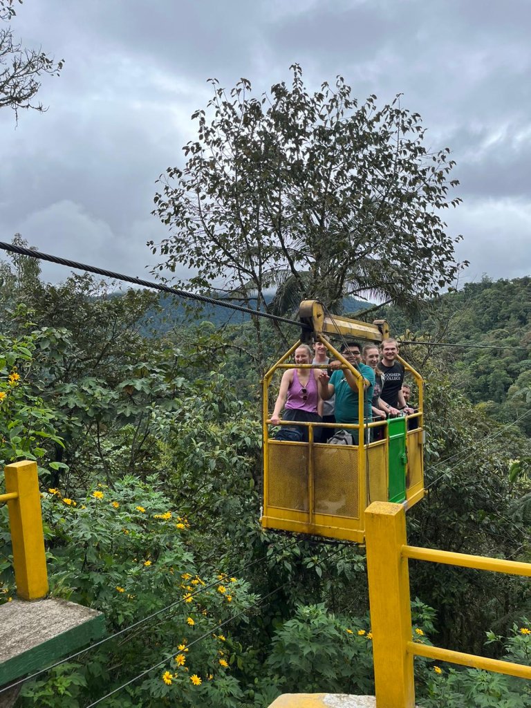 People riding in a yellow open cable car over a green forested valley.