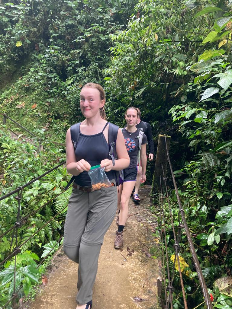 Two smiling hikers walking up a dirt trail through green forest.