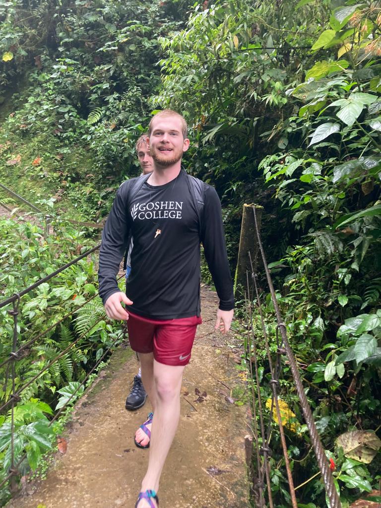 A smiling young man walking down a jungle trail surrounded by greenery.