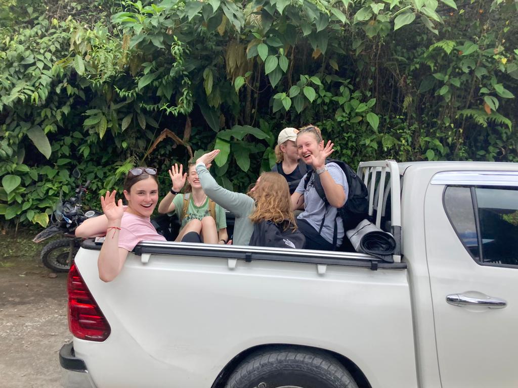 Young people waving from the bed of a silver pickup truck surrounded by green trees.