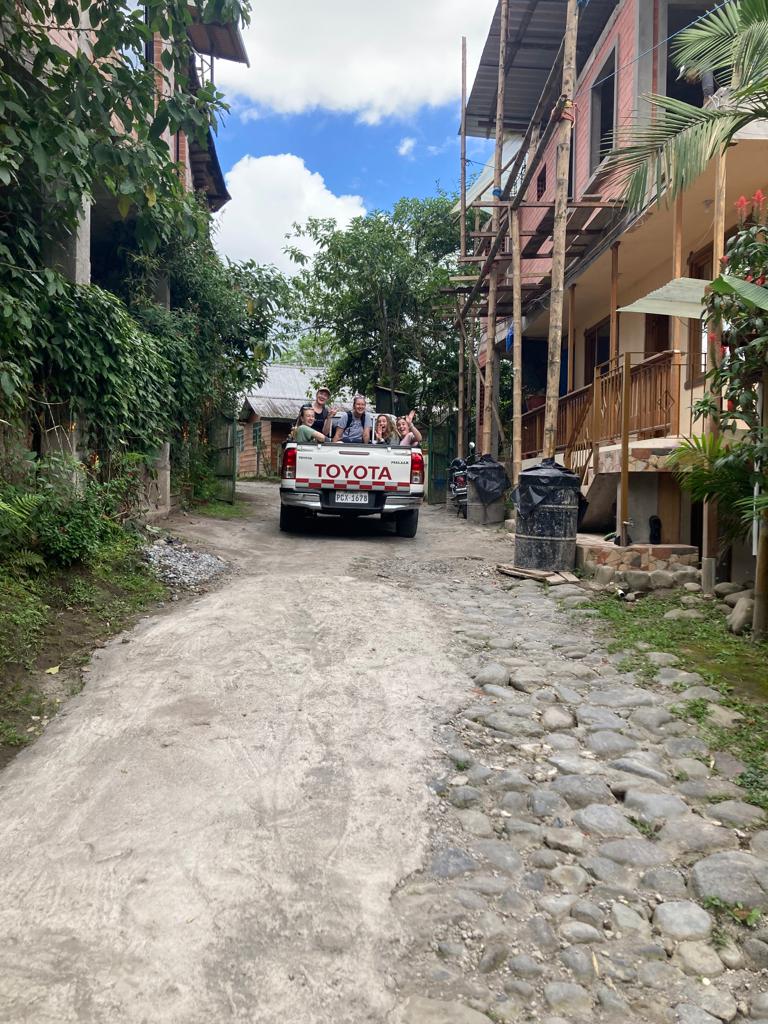 A narrow cobblestone village street lined with buildings, with a truck driving in the distance.